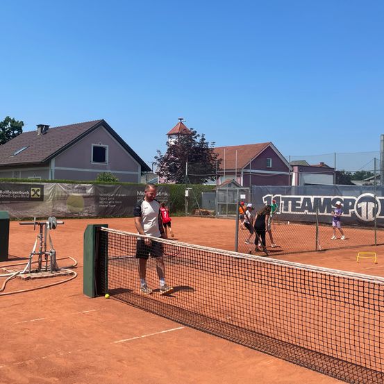 Tennisspieler trainieren auf einem Platz mit Netz, umgeben von Häusern und einem Zaun. Ein Mann steht in der Nähe des Netzes, und andere spielen.