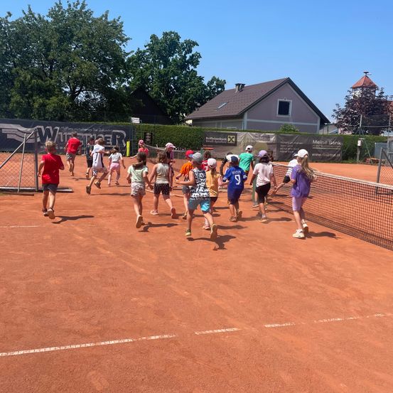Eine Gruppe von Kindern läuft über einen Tennisplatz, einige tragen Mützen und Turnschuhe. Der Platz ist eingezäunt, mit einem Haus und Bäumen im Hintergrund.