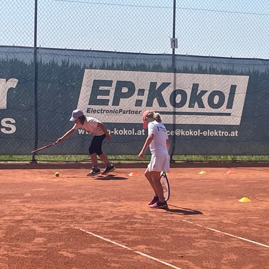 Zwei Tennisspieler auf einem Sandplatz, einer schlägt mit einem Schläger einen Ball, der andere bereitet sich auf den Schlag vor. Ein Zaun mit EP:KokoI-Werbung im Hintergrund.