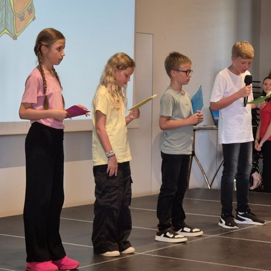 Vier Kinder stehen auf der Bühne, halten Papiere und Mikrofone und lesen davon. Die Bühne hat einen grauen Boden. Hinter ihnen ist eine Projektionsleinwand zu sehen.