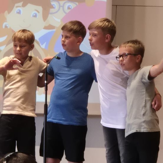 Vier Jungen stehen vor einem Mikrofon, vor einem farbenfrohen animierten Hintergrund. Sie sind alle lässig gekleidet.