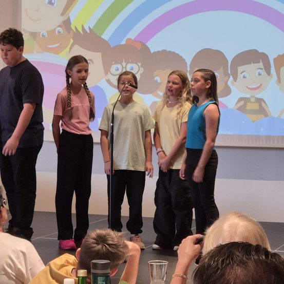 Eine Gruppe von Kindern und ein Junge stehen vor einer Leinwand mit einem Regenbogen. Sie scheinen eine Aufführung zu geben. Eine Person hält den Moment mit einer Kamera fest.
