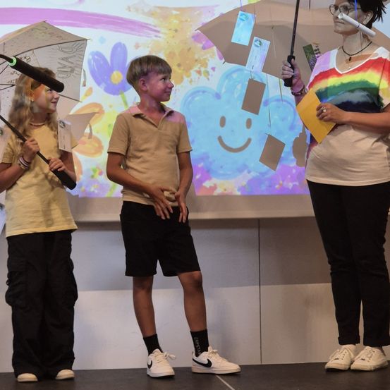 Drei Kinder stehen auf einer Bühne und halten Regenschirme. Ein Mädchen hält ein Mikrofon, ein Junge sieht ernst aus und eine Frau lächelt. Im Hintergrund ist ein farbenfrohes Gemälde zu sehen.