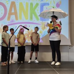 Fünf Kinder stehen mit einem Schirm und einem Mikrofon auf der Bühne, hinter ihnen steht auf einer Leinwand Danke geschrieben.