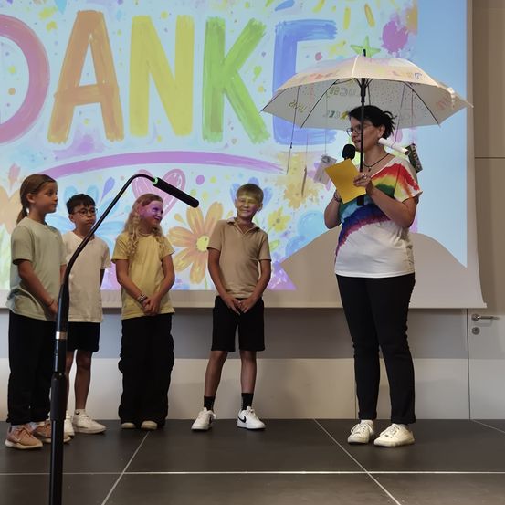 Fünf Kinder stehen mit einem Schirm und einem Mikrofon auf der Bühne, hinter ihnen steht auf einer Leinwand Danke geschrieben.