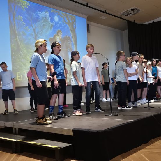 Eine Gruppe von Kindern steht mit Mikrofonen auf der Bühne. Sie stehen wahrscheinlich auf. Ein Beamer zeigt ein Bild hinter ihnen.