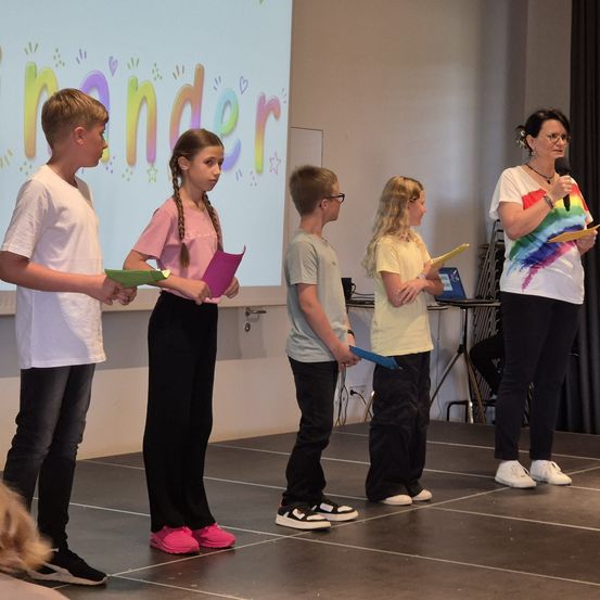 Eine Gruppe von Kindern steht mit einer Frau, die ein Mikrofon hält, auf der Bühne. Sie halten Papiere und nehmen an einer Schulveranstaltung teil.