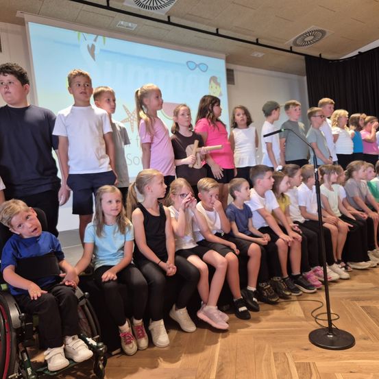 Eine Gruppe von Kindern sitzt auf einer Bühne mit einem Mikrofon davor, einige stehen dahinter, ein Kind im Rollstuhl, einige tragen Brillen, Turnschuhe und Armbänder.