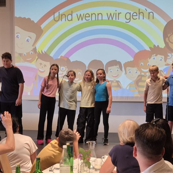 Eine Gruppe von Kindern tritt mit einem Regenbogenhintergrund auf der Bühne auf. Sie stehen vor einer Projektionsleinwand mit Cartoon-Kindern. Einige Erwachsene stehen hinter ihnen, andere sehen dem Publikum zu.