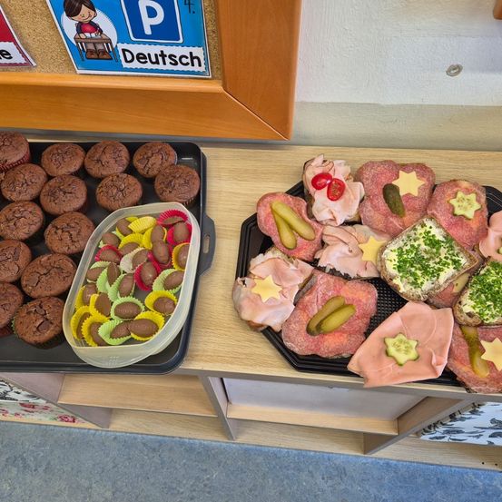 Zwei Tabletts mit Essen auf einem Tisch, ein Tablett mit Schokoladenmuffins und Schokoladen, das andere mit Sandwiches mit Schinken, Gurken und Käse.