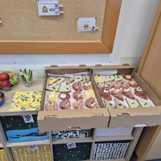 Eine Pappschachtel zeigt eine Vielzahl von Sandwiches und Snacks auf einem Holzregal. Es gibt Obst und Gemüse in der Nähe. Eine Pinnwand ist an der Wand angebracht.