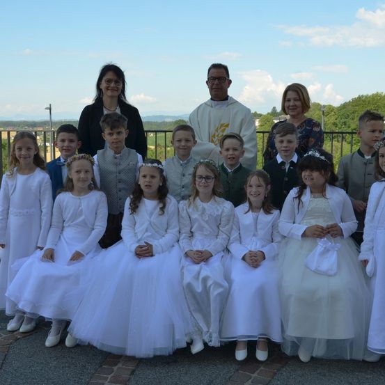 Eine Gruppe von Kindern und Erwachsenen steht draußen, wahrscheinlich für ein Foto. Sie sind in Weiß gekleidet, möglicherweise für eine Zeremonie. Hinter ihnen steht ein Priester, und es gibt Zäune und Bäume im Hintergrund.