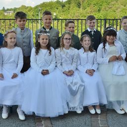 Eine Gruppe von Kindern, die in Weiß gekleidet sind, einige mit Brille, sitzen zusammen vor einem Metallgeländer und lächeln für ein Foto. Bäume und ein Haus sind im Hintergrund.