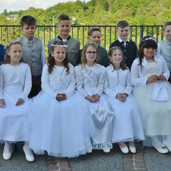 Eine Gruppe von Kindern, die in Weiß gekleidet sind, einige mit Brille, sitzen zusammen vor einem Metallgeländer und lächeln für ein Foto. Bäume und ein Haus sind im Hintergrund.