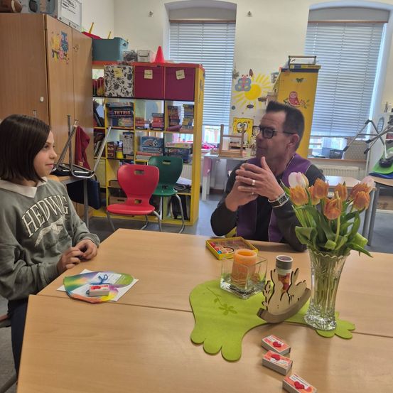 Ein Klassenzimmer mit einem Mann und einem Mädchen, die an einem Tisch sitzen, mit Blumen und Spielzeug. Der Mann trägt eine Brille und spricht, während das Mädchen aufmerksam zuhört.
