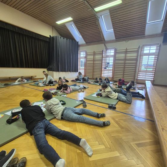 Eine Gruppe von Kindern liegt auf Matten auf dem Boden und schreibt in Notizbücher. Sie befinden sich in einem Klassenzimmer mit Holzdecken und Fenstern.