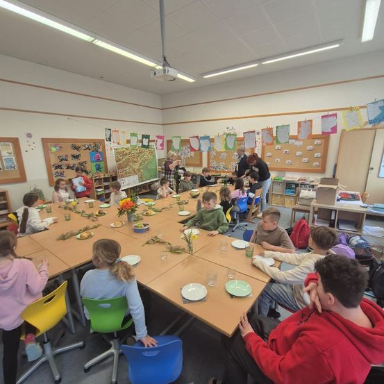 Ein Klassenzimmer mit Kindern, die an langen Tischen sitzen und essen. Ein Beamer hängt an der Decke.