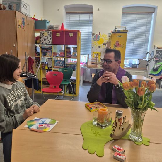 Ein Mann und ein junges Mädchen sitzen sich an einem Tisch gegenüber. Der Tisch hat eine Vase mit Tulpen, eine Kerze und einige kleine Gegenstände. Sie befinden sich in einem Raum mit mehreren Stühlen, Regalen und einem Fenster.