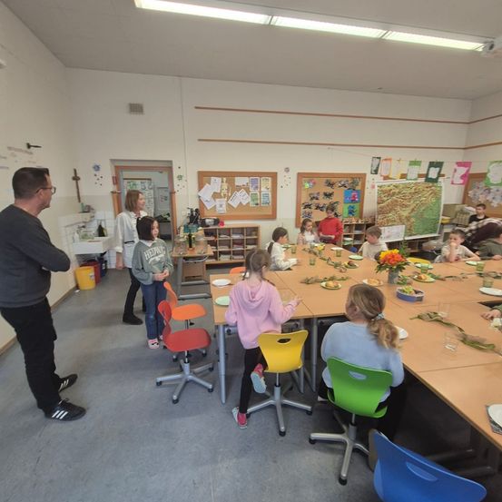 Mehrere Kinder sitzen um einen Tisch herum und essen. Ein erwachsener Mann steht in der Nähe und einige Erwachsene stehen und unterhalten sich. Der Raum hat weiße Wände, Anschlagtafeln und verschiedene Dekorationen.