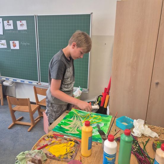 Ein Junge malt auf einer Leinwand in einem Klassenzimmer, mit einer grünen Tafel hinter ihm und verschiedenen Malutensilien auf dem Tisch.