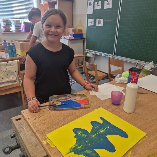 Ein junges Mädchen in einem schwarzen Shirt steht in einem Klassenzimmer und lächelt. Es hält eine Palette mit Farbe und einen Pinsel. Ein gelbes Papier mit einem blauen Fisch liegt auf dem Tisch.