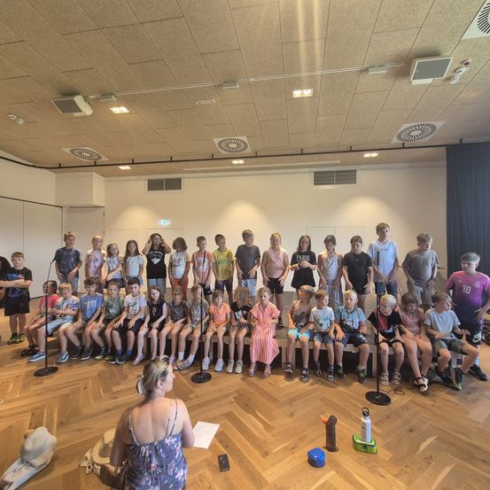 Eine Gruppe von Kindern und einer Frau versammelt sich in einem Innenraum. Sie sitzen auf einer Bühne mit Mikrofonen und nehmen wahrscheinlich an einer Chorprobe teil.