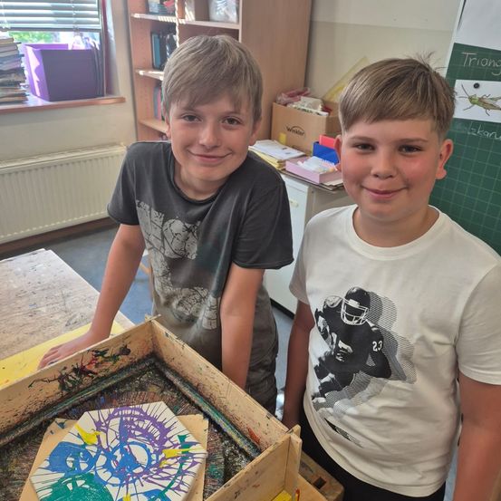 Zwei junge Jungen lächeln und stehen neben einer Kiste voller bunter, verspritzter Kunst. Ein Junge trägt ein graues T-Shirt mit Grafikdesign, der andere ein weißes T-Shirt mit einem Footballspieler-Design. Dahinter befindet sich ein Holzregal mit Büchern und einer Tafel.
