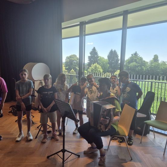 In einem Raum mit Holzboden stehen und sitzen eine Gruppe von Kindern und einem Erwachsenen um einen Notenständer herum und spielen Instrumente. Der Erwachsene spielt Akkordeon, während die Kinder singen. Dahinter befindet sich eine Glaswand mit Blick auf Bäume und Gras.