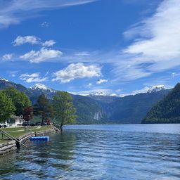 Eine ruhige Seeansicht mit Bergen im Hintergrund, Bäumen und einem klaren blauen Himmel. Es gibt Autos in der Nähe des Ufers und ein kleines blaues Objekt schwimmt im Wasser.