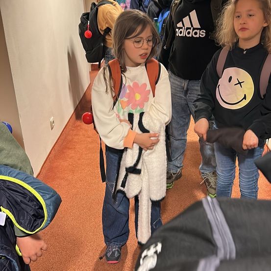 Ein Mädchen mit Brille hält einen weißen Plüschtier in einem Flur mit anderen Kindern. Ein Adidas-Logo ist auf dem Shirt einer Person zu sehen.