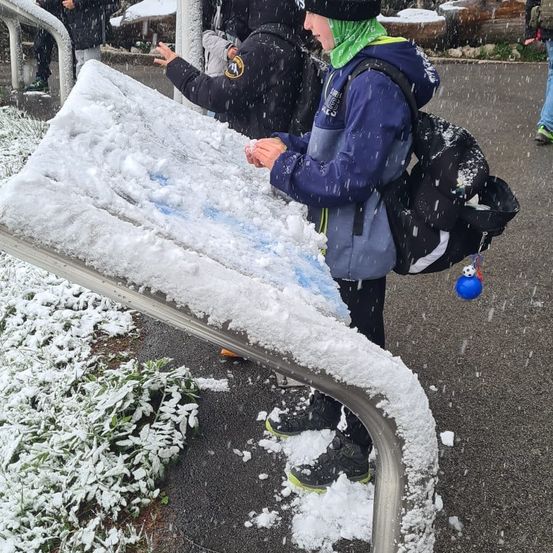 Zwei Personen stehen auf einer schneebedeckten Karte, tragen Winterkleidung und haben Rucksäcke. Sie scheinen die Karte zu betrachten, während Schnee um sie herum fällt.