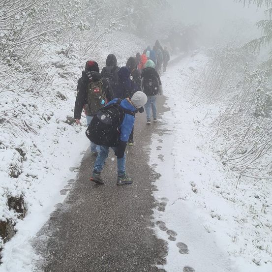 Eine Gruppe von Menschen wandert im Schnee und trägt Winterkleidung und Rucksäcke. Der Weg ist schneebedeckt, und sie hinterlassen Fußabdrücke.