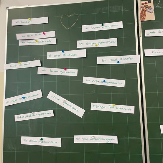 Eine grüne Tafel mit Haftnotizen und Pins zeigt verschiedene Sätze auf Deutsch. Bemerkenswerte Sätze sind 'Wir spielen zusammen,' 'Wir singen zusammen' und 'Wir helfen uns gegenseitig.' Die Tafel hat auch ein Herz gezeichnet mit einem Faden.