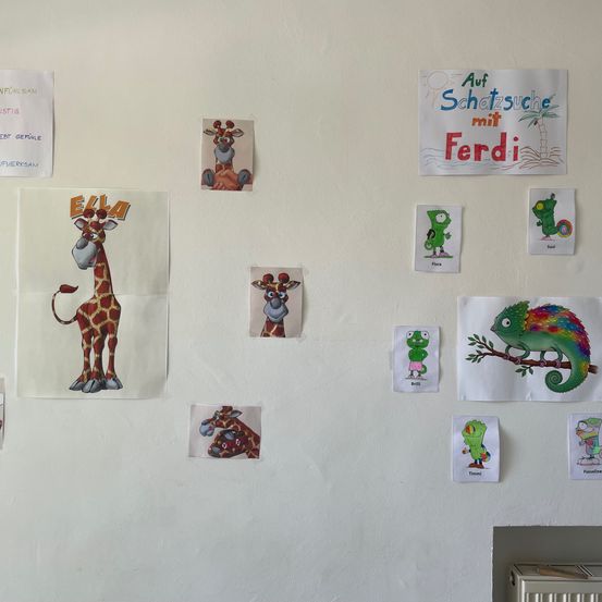 Eine weiße Wand mit mehreren Bildern und Schildern. Die Bilder zeigen eine Giraffe, einen Chamäleon und ein Poster mit dem Text 'Auf Schatzsuche mit Ferdi'. Die Schilder sind 'Ella', 'Flora', 'Soil' und 'Timmi'. Unten rechts befindet sich eine Heizung.
