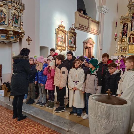 Eine Gruppe von Kindern in verschiedenen Mänteln und Mützen steht in einer Kirche, einige tragen weiße Gewänder. Eine Frau in einem schwarzen Mantel steht vor ihnen.