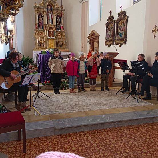 In einer Kirche steht eine Gruppe von Kindern auf der Bühne, während ein Mann eine Gitarre spielt. Dahinter steht eine Kanzel mit einem Mikrofon. An der Wand befinden sich religiöse Ikonen und Kreuze.