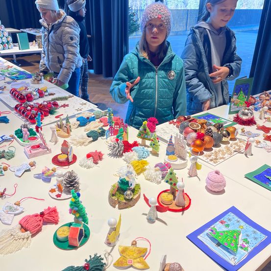 Drei Mädchen stehen vor einem Tisch voller handgefertigter Weihnachtsdekorationen, wie Ornamente, Girlanden und kleine Figuren. Ein Mädchen zeigt auf eine Ausstellung.