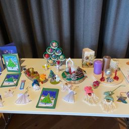 Ein Tisch zeigt verschiedene Weihnachtsdekorationen, darunter Kiefernzapfen, Kerzen, Ornamente und handgefertigte Bastelarbeiten. Das Ambiente ist schlicht mit hellem Hintergrund.
