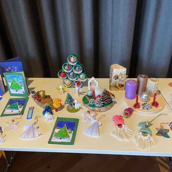 Ein Tisch zeigt verschiedene Weihnachtsdekorationen, darunter Kiefernzapfen, Kerzen, Ornamente und handgefertigte Bastelarbeiten. Das Ambiente ist schlicht mit hellem Hintergrund.