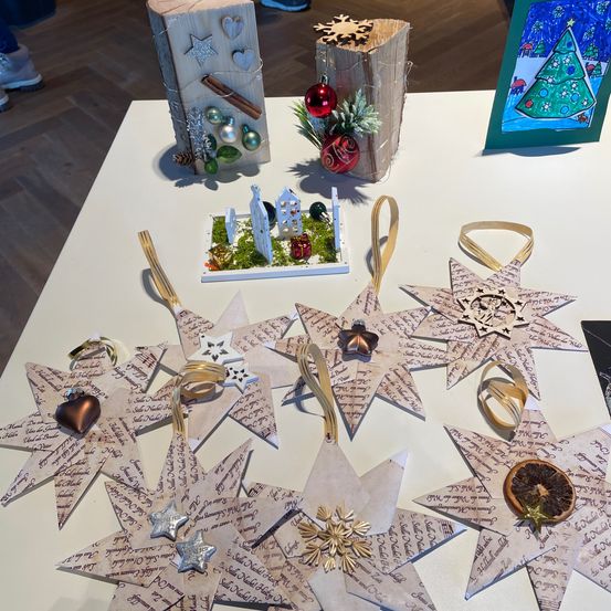 Ein Tisch zeigt handgefertigte Weihnachtsdekorationen. Sternförmige Ornamente und Mini-Holzhäuser gehören zu den ausgestellten Gegenständen. Rechts ist ein Rahmen mit einer Weihnachtsbaum-Malerei.