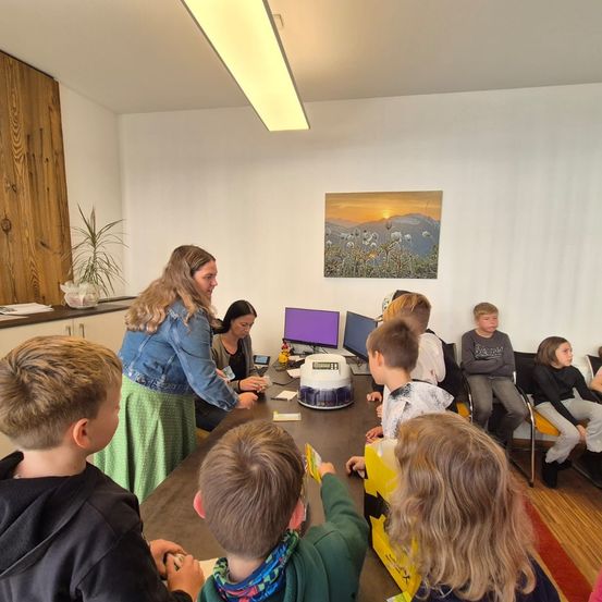 Eine Gruppe von Kindern und Erwachsenen sitzt um einen Tisch mit einer Maschine herum. Eine Frau steht hinter dem Tisch und spricht mit den Kindern. An der Wand hängt ein Bilderrahmen.