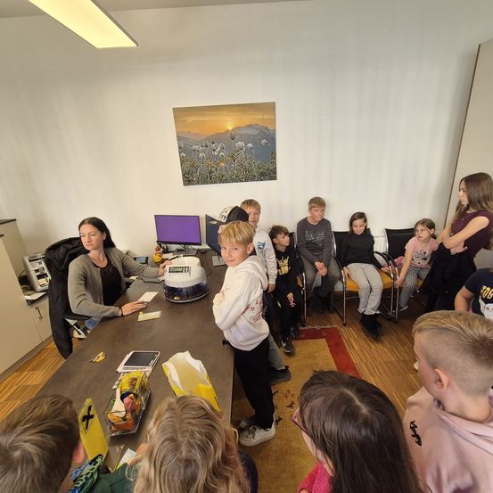 Eine Gruppe von Kindern und Erwachsenen ist um einen Tisch herum versammelt. Eine Frau sitzt mit verschiedenen Gegenständen auf dem Tisch. Der Raum hat weiße Wände und Holzböden.