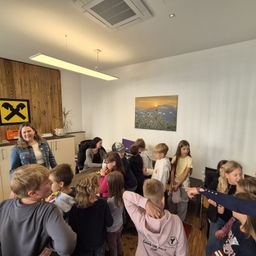 Eine Gruppe von Kindern und Erwachsenen versammelt sich in einem Raum mit Holzwand, Bild und Tisch. Die Kinder schauen auf etwas auf dem Tisch, während die Erwachsenen sie beobachten.