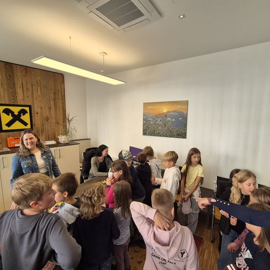 Eine Gruppe von Kindern und Erwachsenen versammelt sich in einem Raum mit Holzwand, Bild und Tisch. Die Kinder schauen auf etwas auf dem Tisch, während die Erwachsenen sie beobachten.