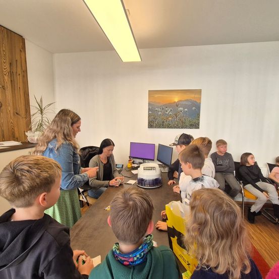 Eine Gruppe von Kindern versammelt sich in einem Klassenzimmer um eine Frau. Sie demonstriert ihnen etwas. Sie sitzen um einen Tisch herum mit Computern und anderen Gegenständen. Ein Topf mit einer Pflanze und ein gerahmtes Bild hängen an der Wand.