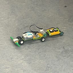 Ein Lego-Fahrzeug mit Rädern und einer Batterie-Einrichtung, verbunden durch Kabel, liegt auf einer grauen Oberfläche.