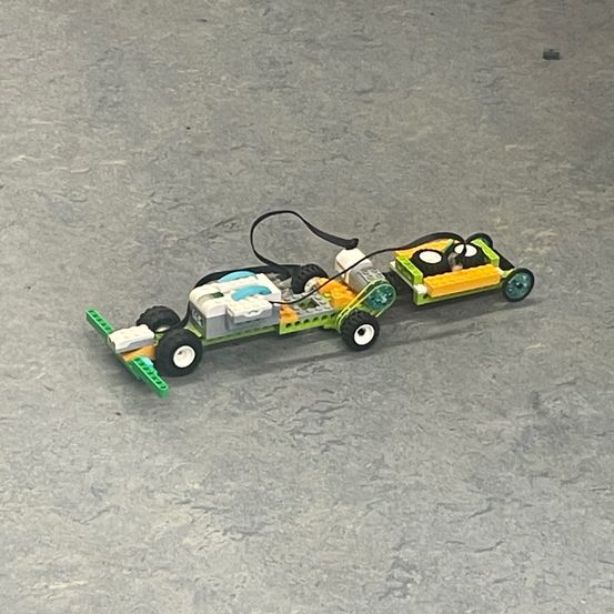 Ein Lego-Fahrzeug mit Rädern und einer Batterie-Einrichtung, verbunden durch Kabel, liegt auf einer grauen Oberfläche.