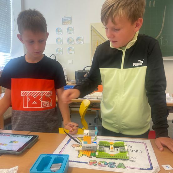 Zwei Jungen im Klassenzimmer, einer trägt ein B&S Style-Shirt und der andere eine Puma-Jacke, konzentrieren sich auf ein Lego-Projekt auf einem Tisch, mit einem Tablet und einem grünen Brett in der Nähe.