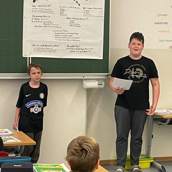 Ein junger Junge in einem schwarzen T-Shirt steht vor einem Klassenzimmer und hält ein Blatt Papier in der Hand. Ein anderer Junge steht hinter ihm in der Nähe eines Schreibtisches mit einem Buch. Hinter ihnen befindet sich eine grüne Tafel mit Aufschriften.