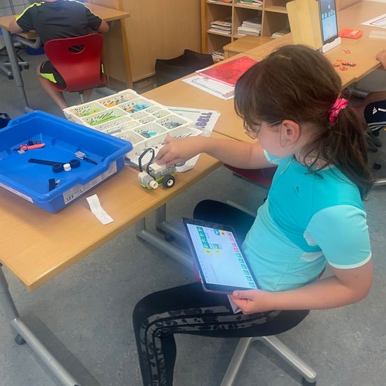 Ein Mädchen sitzt im Klassenzimmer und hält ein Tablet in der Hand, während es an einem Projekt mit einem Roboter arbeitet. Vor ihr stehen Aufbewahrungsboxen mit Teilen und Werkzeugen.